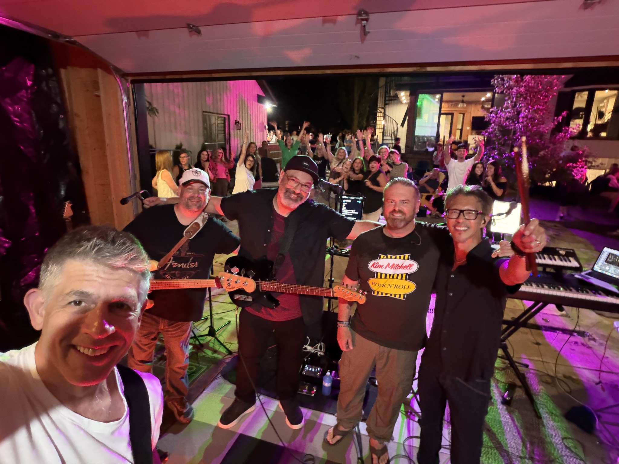 A band selfie with instruments, colourful stage lights, and a cheering crowd behind them.